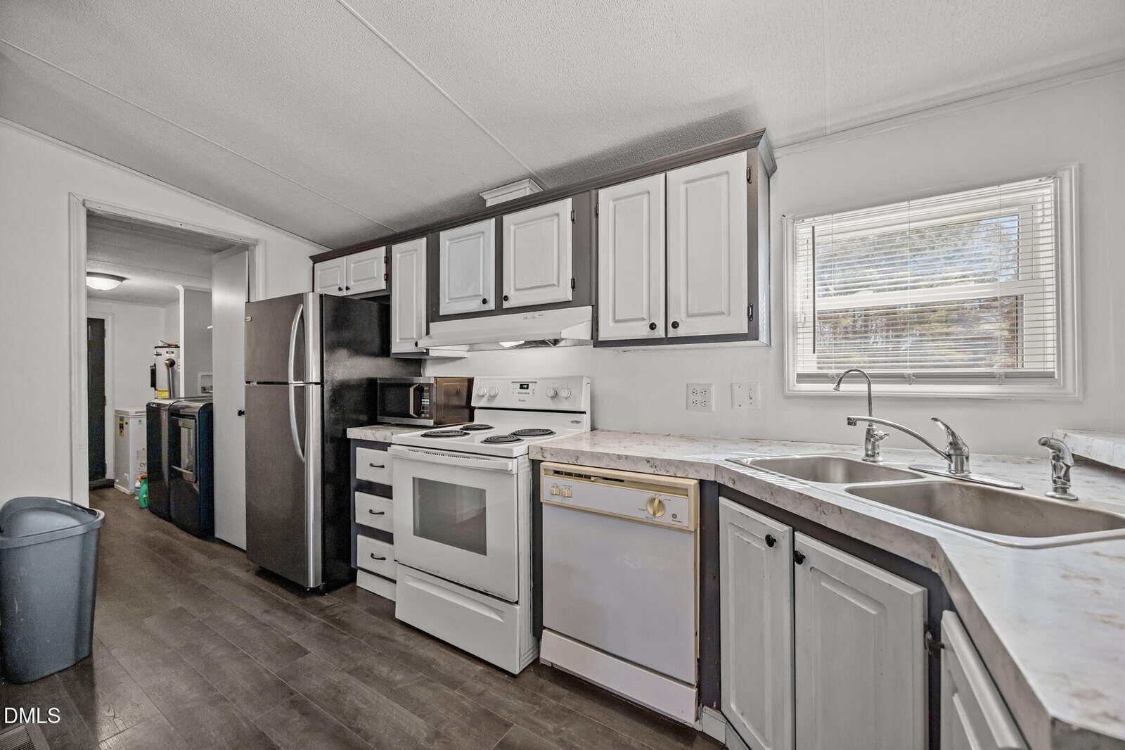 A kitchen with white and dark cabinets, white appliances, a stainless steel refrigerator, double sink under a window, and dark wood floors. A trash bin and laundry area are visible through an open doorway.