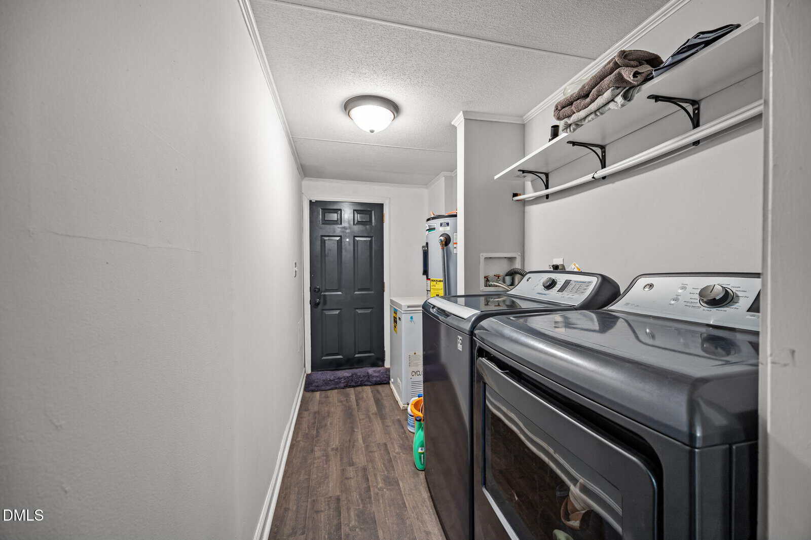 A narrow laundry room with a dark washer and dryer on the right, shelves above holding towels and supplies, white walls, wood flooring, and a black door at the end of the hallway.