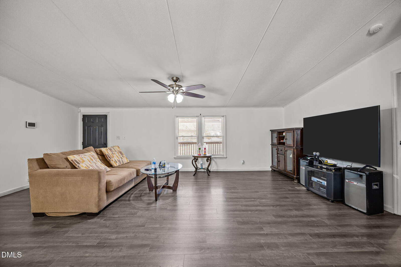 A spacious living room with wood flooring, a tan sofa with patterned pillows, a glass coffee table, a large flat-screen TV on a dark entertainment center, and a ceiling fan with lights. White walls and a window provide natural light.