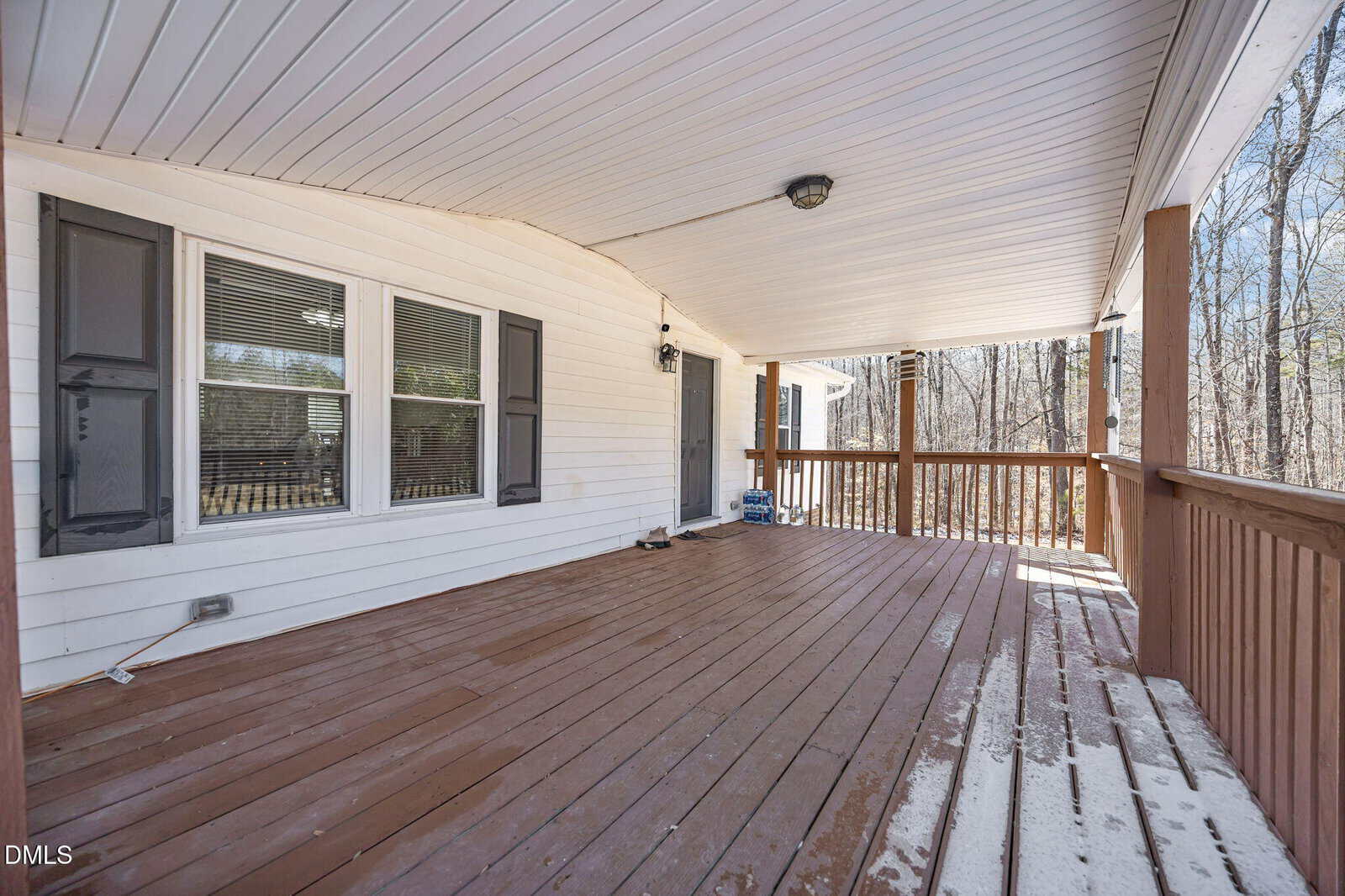 A spacious covered wooden porch with railings, white siding, three windows with dark shutters, and a wooded area visible in the background. Some sunlight and shadows are cast on the floor.