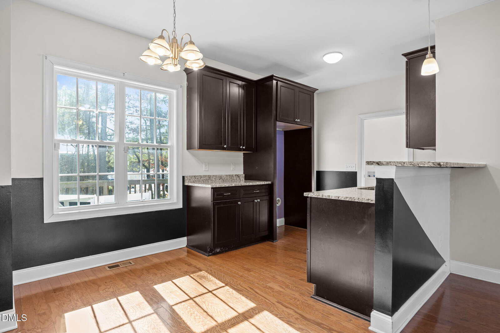 Bright kitchen with dark wood cabinets, granite countertops, and hardwood floors. Large window lets in natural light. Pendant and chandelier lighting hang from the ceiling. Walls are two-toned in white and dark gray.