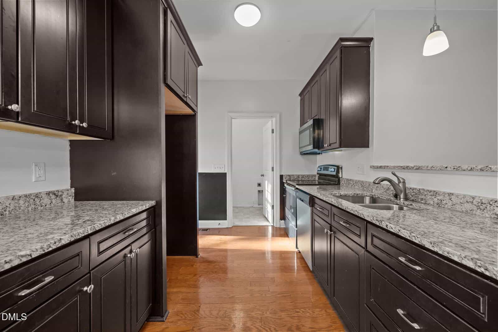 A modern kitchen with dark wood cabinets, granite countertops, stainless steel appliances, double sink, and hardwood floors. The space is well-lit with ceiling and pendant lights.