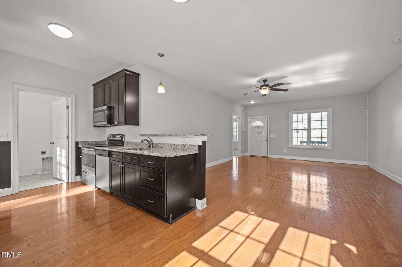 Open-plan kitchen and living area with wood flooring, dark cabinets, granite countertops, stainless steel appliances, large window, ceiling fan, and white walls. Natural light brightens the spacious, modern interior.