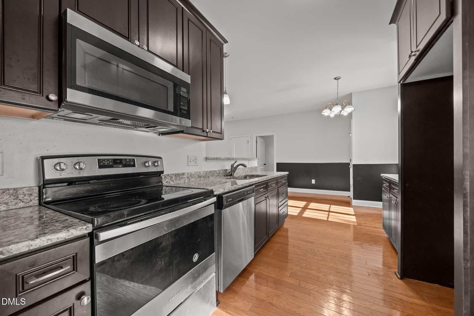 Modern kitchen with dark cabinets, stainless steel appliances, granite countertops, and hardwood floors, open to a bright dining area with large windows and a hanging light fixture.