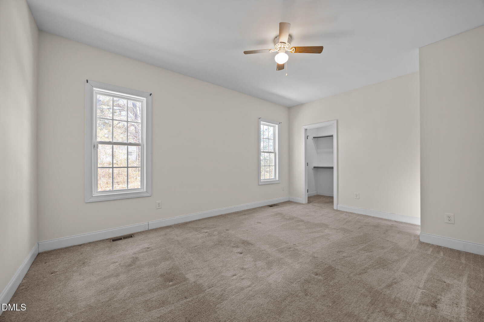 Empty bedroom with beige carpet, white walls, two windows, small ceiling fan with light, and an open closet. Natural light fills the space from the windows on the left wall.
