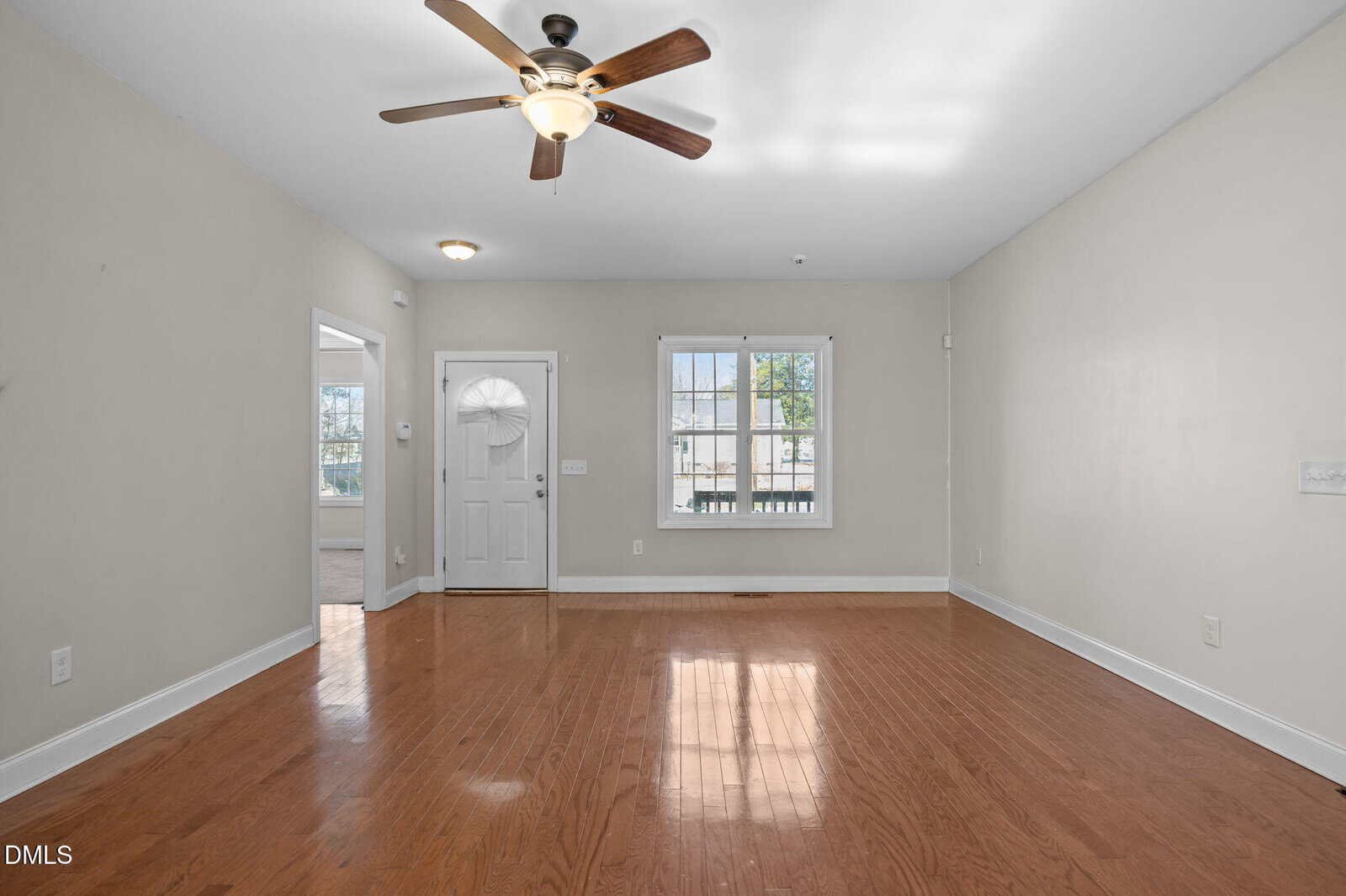 Bright, empty living room with light beige walls, hardwood floors, a ceiling fan with light, two windows, and a white front door. Sunlight streams through the windows, creating a welcoming atmosphere.