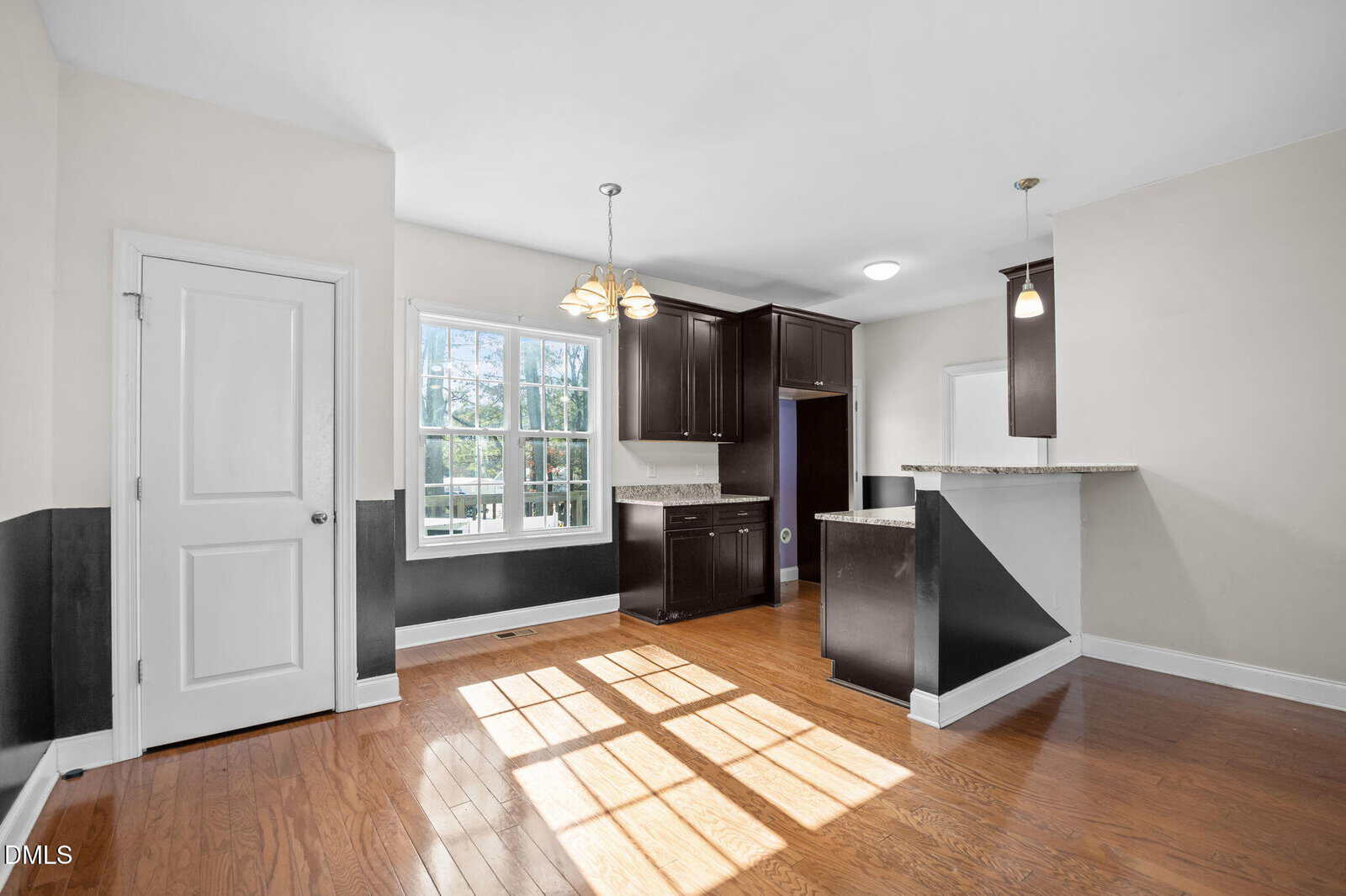 Bright kitchen with dark wood cabinets, granite countertops, pendant lights, hardwood floors, a window letting in natural light, and a door to the left. Walls are painted white with a dark accent stripe.