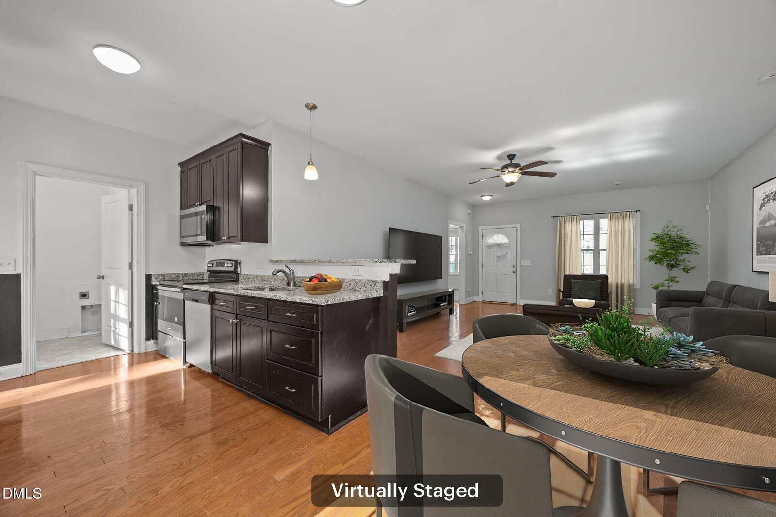 Open-concept living space with wood flooring, modern kitchen with dark cabinets, dining table with decor, and living area featuring a sofa, TV, and ceiling fan. Image labeled "Virtually Staged.