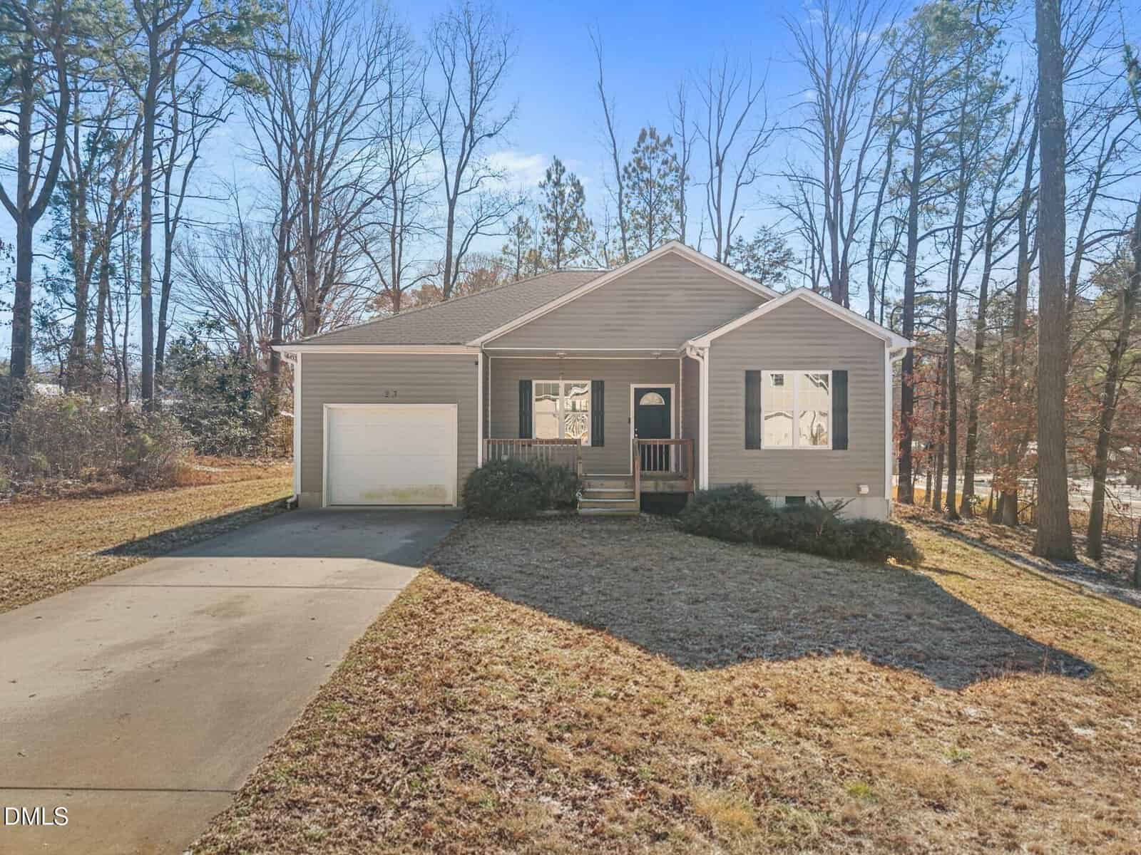 A single-story beige house with an attached garage, small front porch, and driveway, surrounded by leafless trees and a grassy yard under a clear blue sky.