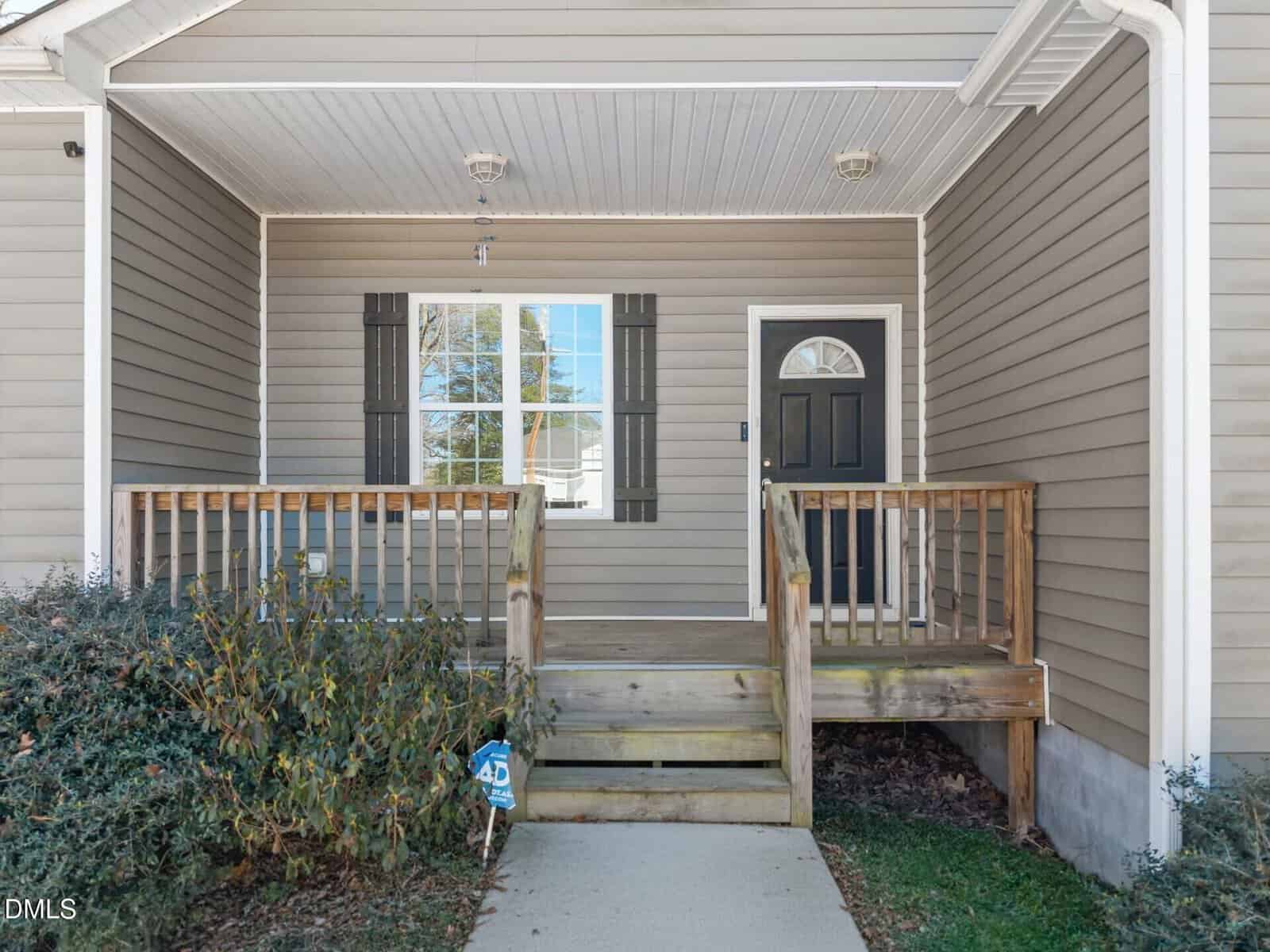A small wooden porch with railings leads to a black front door on a beige house with white trim and a large window with dark shutters. Shrubs are on the left side of the entrance.