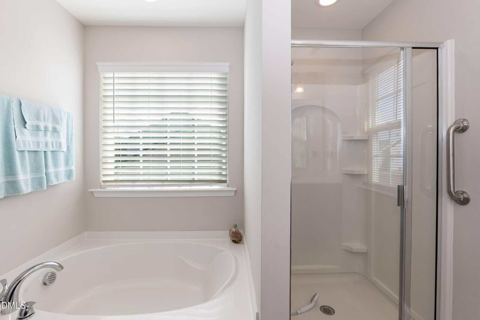 A bathroom with a white bathtub next to a window with blinds, a glass-enclosed shower, and light-colored walls. Blue towels hang on the wall above the tub.