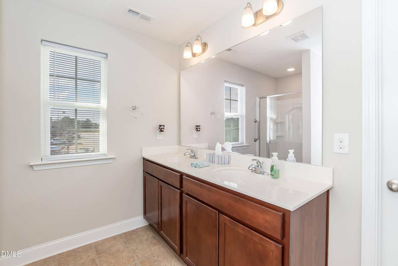 A bright bathroom with a double sink vanity, large mirror, two wall lights, a window, towel ring, and a glimpse of a shower in the background. The cabinets are wooden with white countertops.