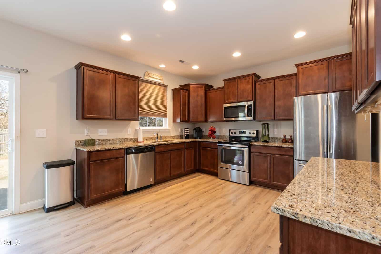 Modern kitchen with wooden cabinets, stainless steel appliances, granite countertops, light wood flooring, and recessed lighting. There is a window above the sink and a sliding glass door to the left.
