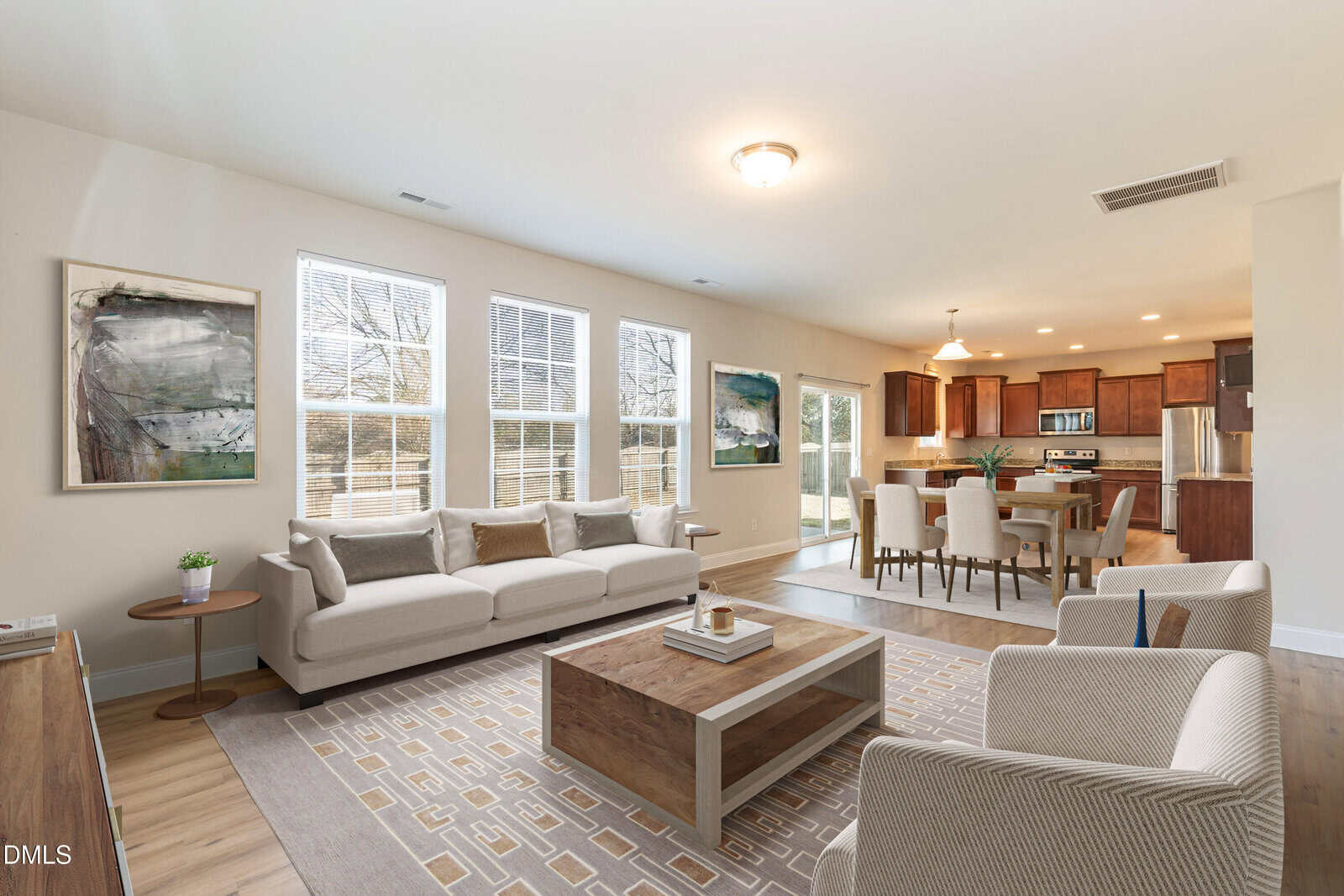 Bright, modern open-plan living and dining area with light walls, large windows, beige sofas, a wood coffee table, dining table with chairs, and a kitchen with wooden cabinets in the background.