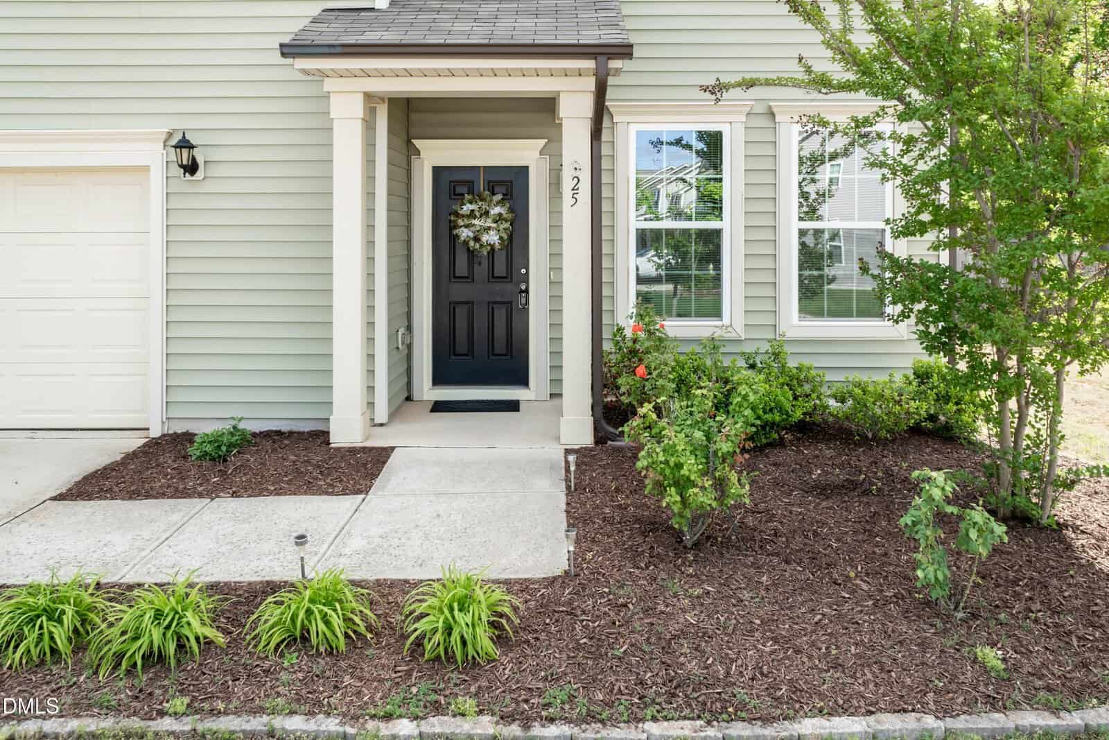 A tidy house entrance with a black front door decorated with a wreath, white trim, light green siding, and landscaped garden beds with shrubs, plants, and mulch.
