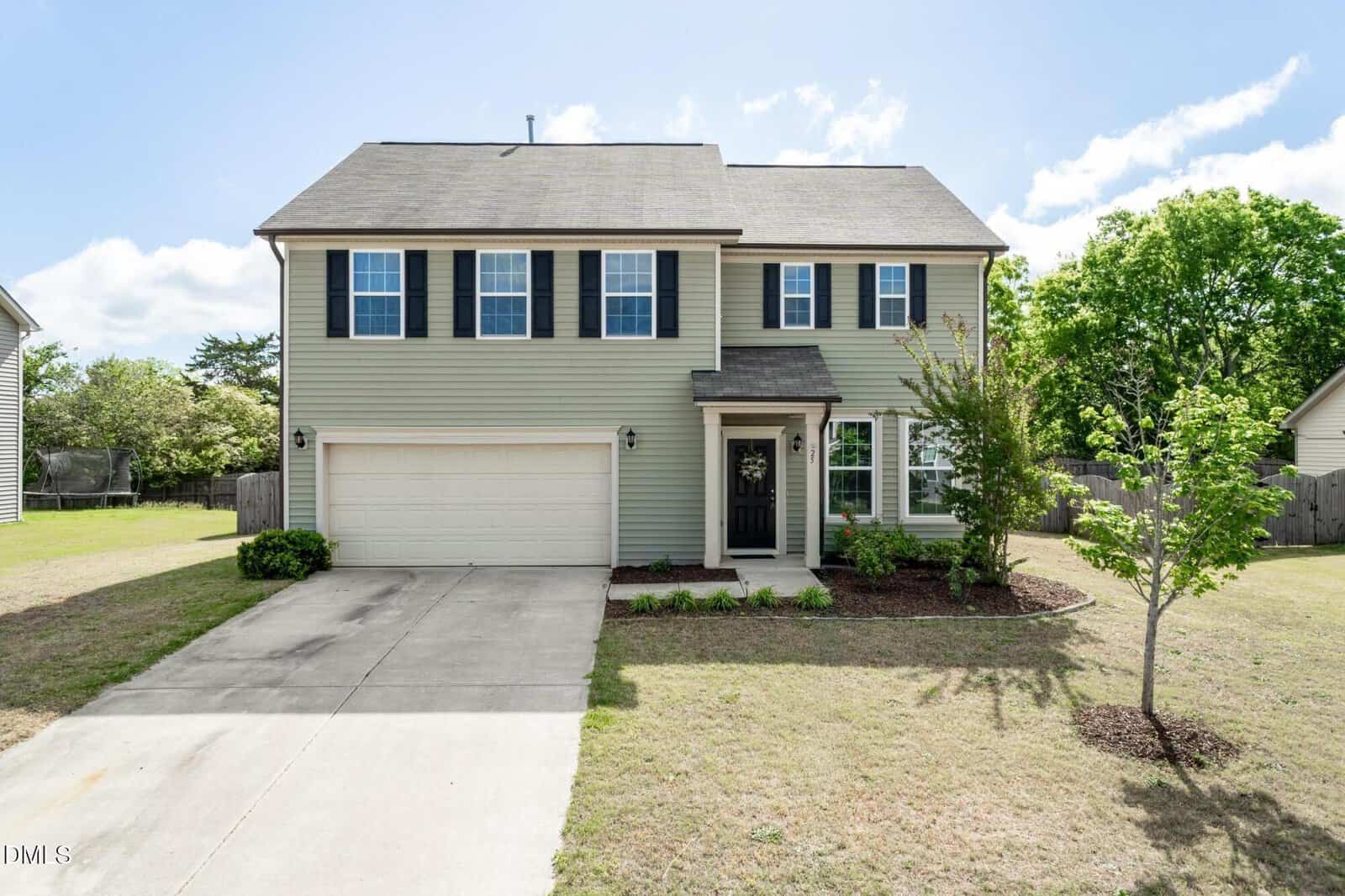 Charming two-story suburban home at 25 Duchess Avenue, Franklinton, with light green siding, black shutters, and a two-car garage. The front yard offers a driveway, small trees, and manicured bushes beneath a blue sky with scattered clouds.