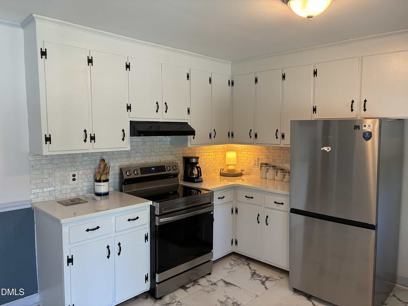 A modern kitchen with white cabinets, stainless steel appliances, marble floor, and a tiled backsplash. Under-cabinet lighting softly illuminates the countertop, which holds a coffee maker, utensils, and storage jars.