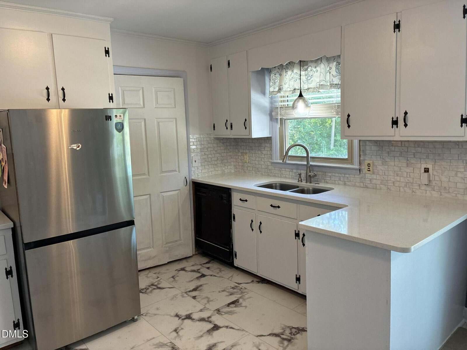 A kitchen with white cabinets, marble-patterned floor, stainless steel fridge, black dishwasher, double sink under a window with a valance, and light subway tile backsplash.