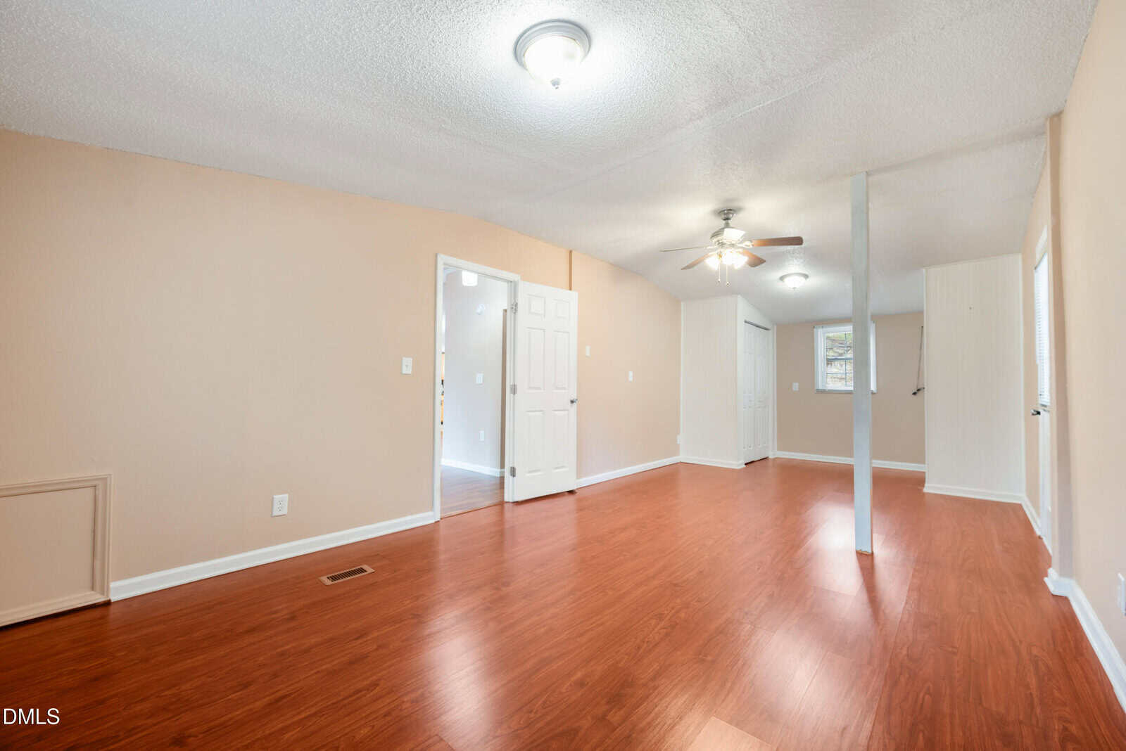 A spacious, empty room with light beige walls, white doors, ceiling lights, a ceiling fan, and reddish-brown wood flooring. There are several doors and a small window letting in natural light.