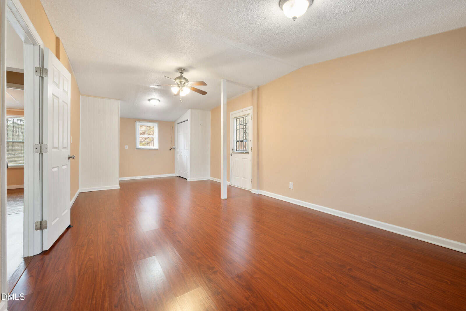 Spacious empty room with light brown walls, wooden floor, ceiling fan, two ceiling lights, white door with glass window, and two windows letting in natural light.