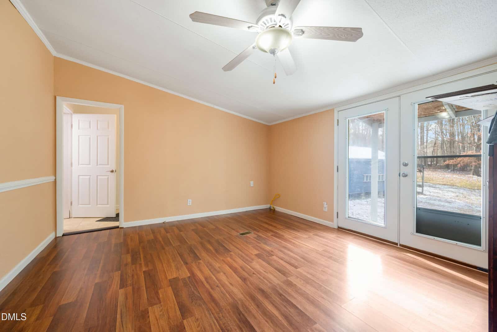 A bright, empty room with light brown walls, wood flooring, a white ceiling fan, double glass doors leading outside, and a single door to another room. Sunlight streams in through the glass doors.
