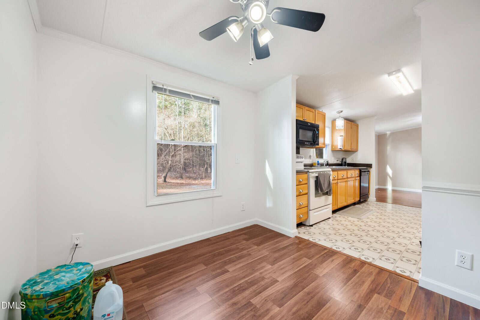 Bright kitchen and dining area with wood floors, white walls, and a window. Kitchen has light wood cabinets, black appliances, and patterned linoleum flooring. Ceiling fan and containers are in the dining space.