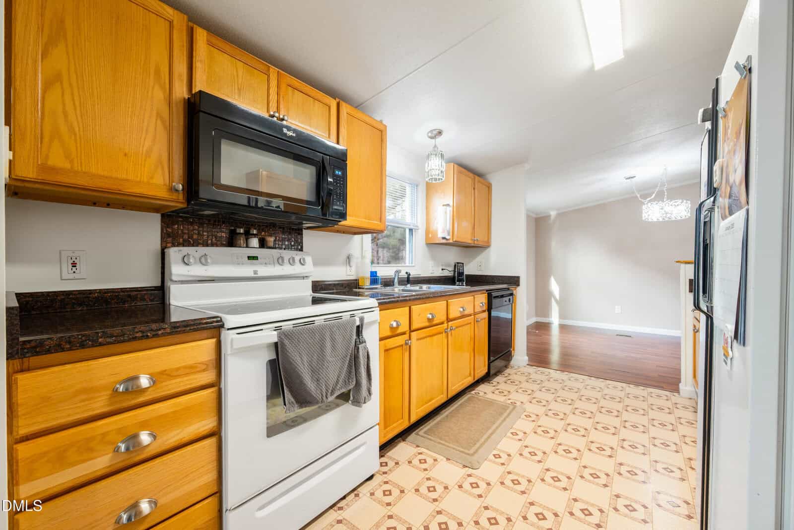 Bright kitchen with light wood cabinets, white stove and microwave, black countertops, patterned floor, and a window over the sink. Adjoining dining area with wood flooring and a modern chandelier.