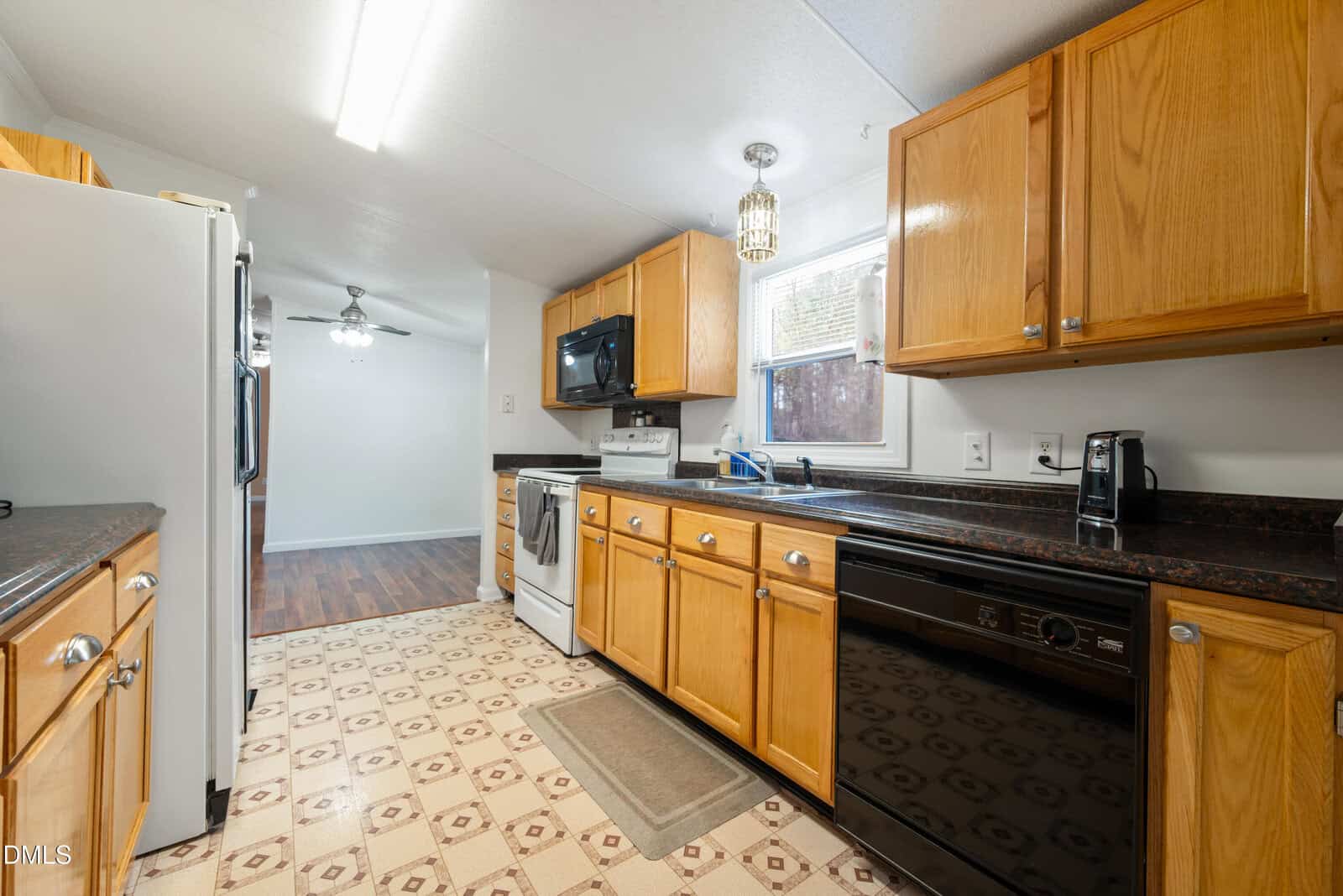 A kitchen with light wood cabinets, black countertops, white appliances, a dishwasher, microwave, and a window above the sink. The floor has a patterned linoleum, and the kitchen opens into a dining area with wood flooring.