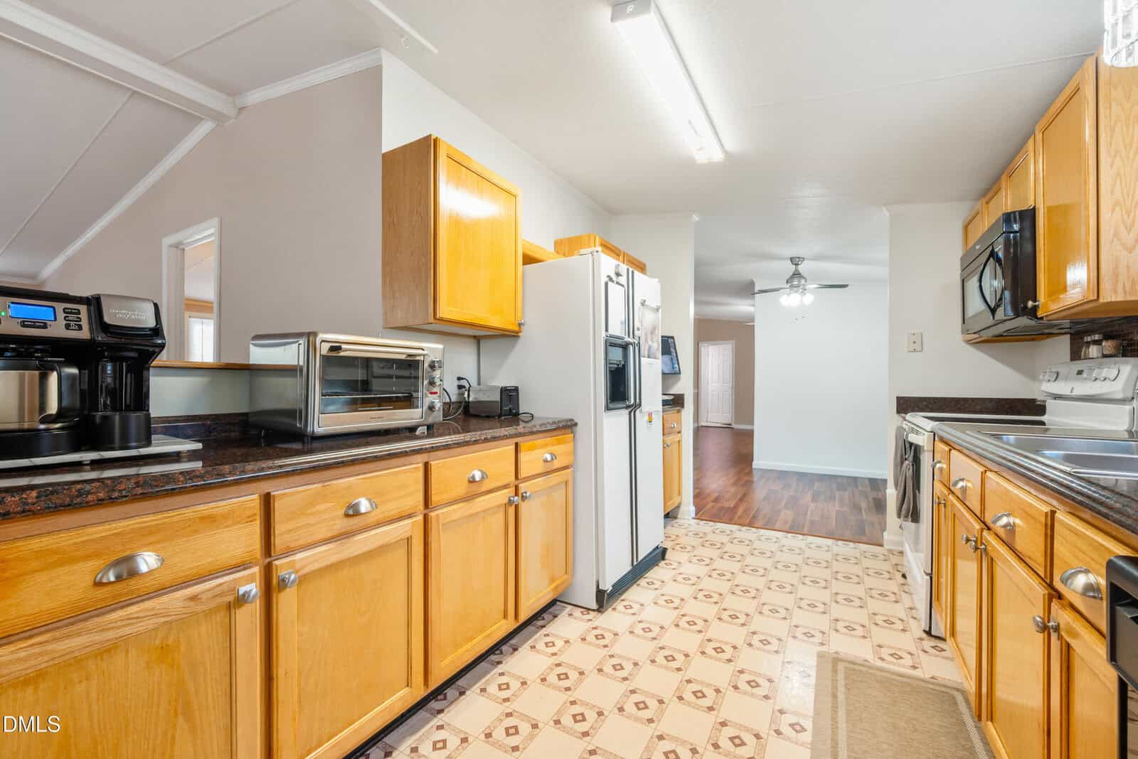 A bright kitchen with light wood cabinets, a white refrigerator, stove, and microwave, small appliances on the counter, patterned floor, and view into a dining area with wood flooring and ceiling fan.