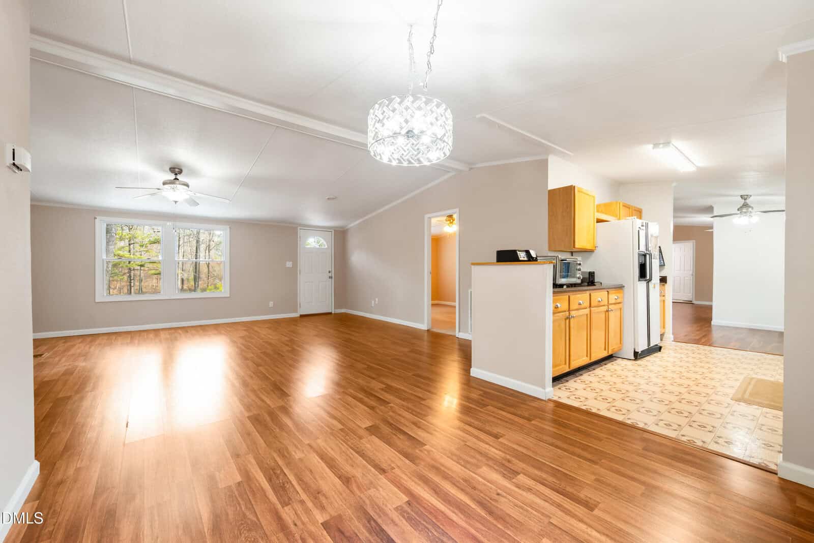 Spacious open-concept living area with wood flooring, large windows, ceiling fans, and an adjacent kitchen featuring wooden cabinets and white appliances. Neutral-colored walls and ample natural light throughout.