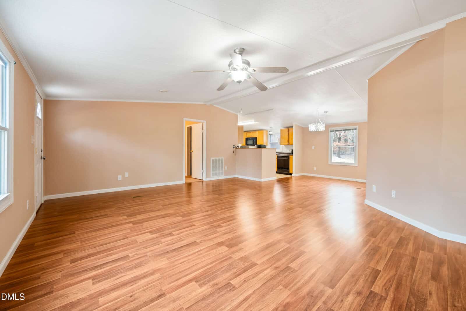Spacious open-concept living area with light brown wood floors, beige walls, a ceiling fan, and a view into a kitchen with white ceilings and wood cabinets. Large windows let in natural light.