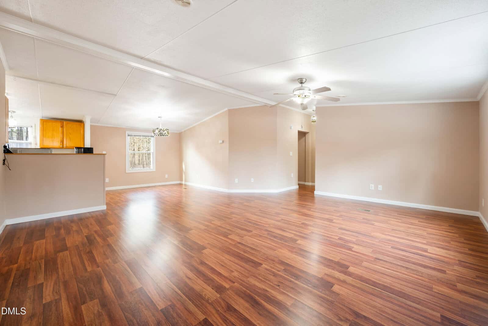 Spacious empty living area with wood flooring, beige walls, white ceiling, two ceiling fans, and a view of the kitchen with wooden cabinets and a window letting in natural light.