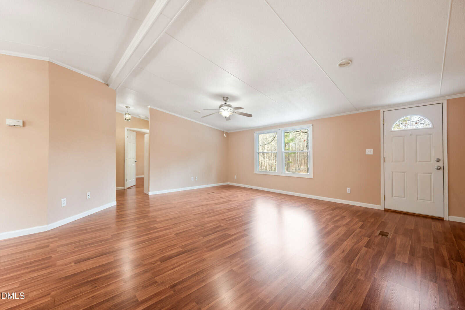 Spacious, empty living room with light brown walls, wood flooring, two windows, a ceiling fan, and a white front door. Natural light comes through the windows, giving the room a bright, airy feel.