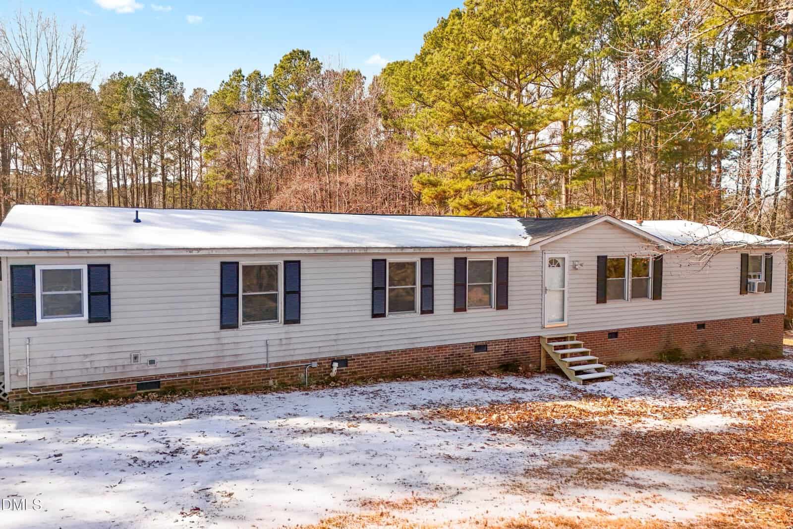 A long, single-story manufactured home with gray siding, blue shutters, and a white door stands on a brick foundation. Pine trees surround the house, and the ground is lightly covered with snow.