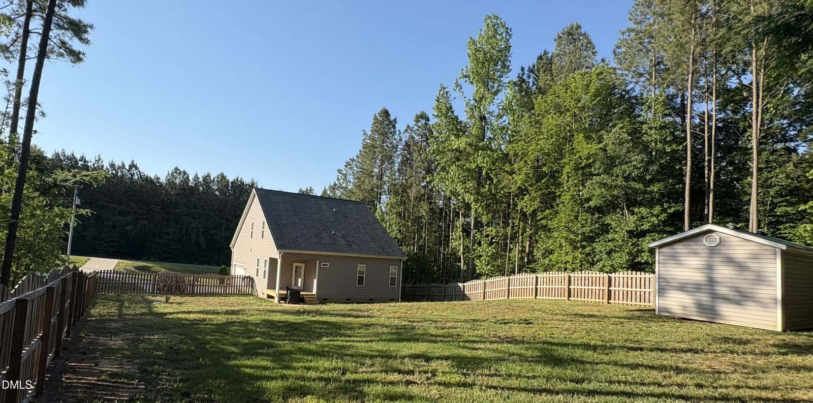 A fenced backyard with a grassy lawn, a small beige house with a sloped roof, a shed, and tall trees in the background under a clear blue sky.