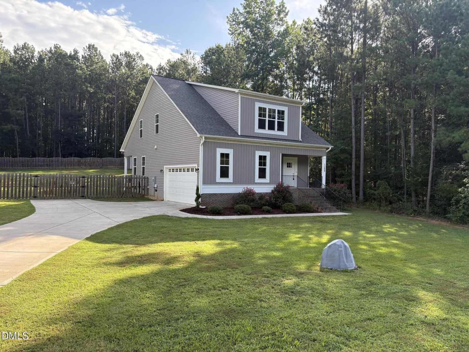 Charming two-story gray house with white trim, located at 1208 Montgomery Road in the heart of Franklinton. Featuring an attached garage, inviting front porch, and a manicured lawn, this home is surrounded by mature trees and a wooden fence. The driveway gracefully curves toward the garage, while a covered object sits on the grass in the foreground.