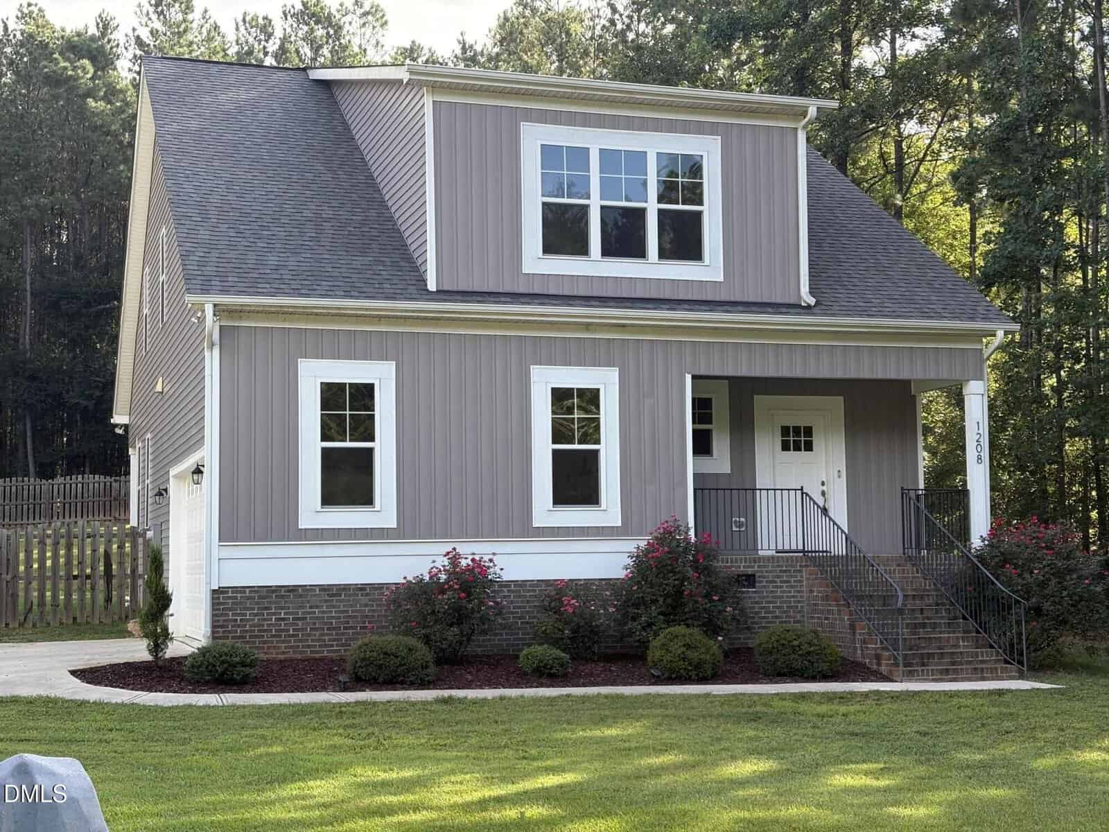 A two-story gray house with white trim, a covered front porch, stairs with a black railing, brick foundation, and landscaped bushes, set against a backdrop of trees and a fenced yard.