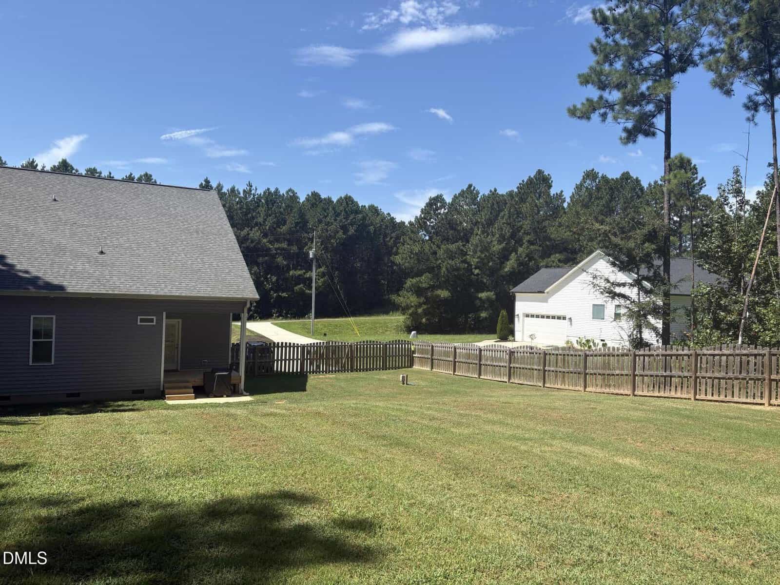 A fenced backyard with a well-kept lawn, part of a gray house on the left, another white house in the background, surrounded by tall trees under a blue sky with some clouds.