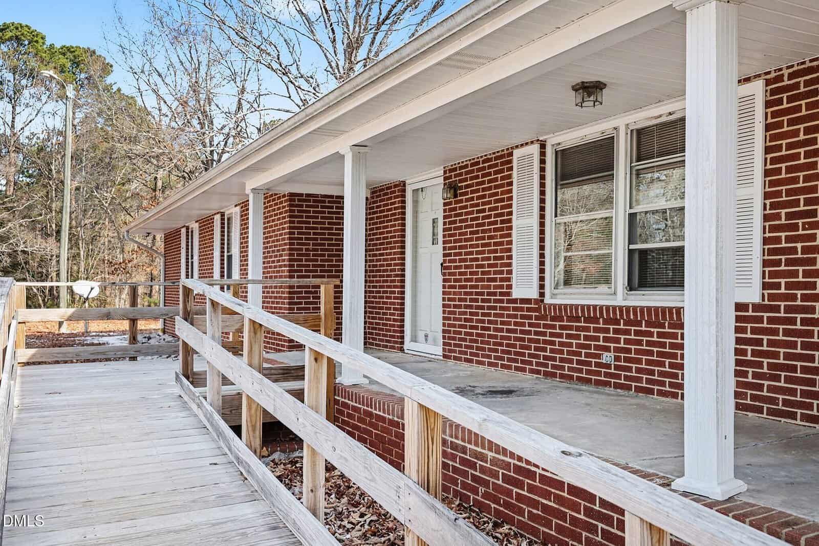 A red brick house with white trim and shutters features a covered front porch and a wooden wheelchair-accessible ramp leading to the entrance. Leafless trees and a yard are visible in the background.