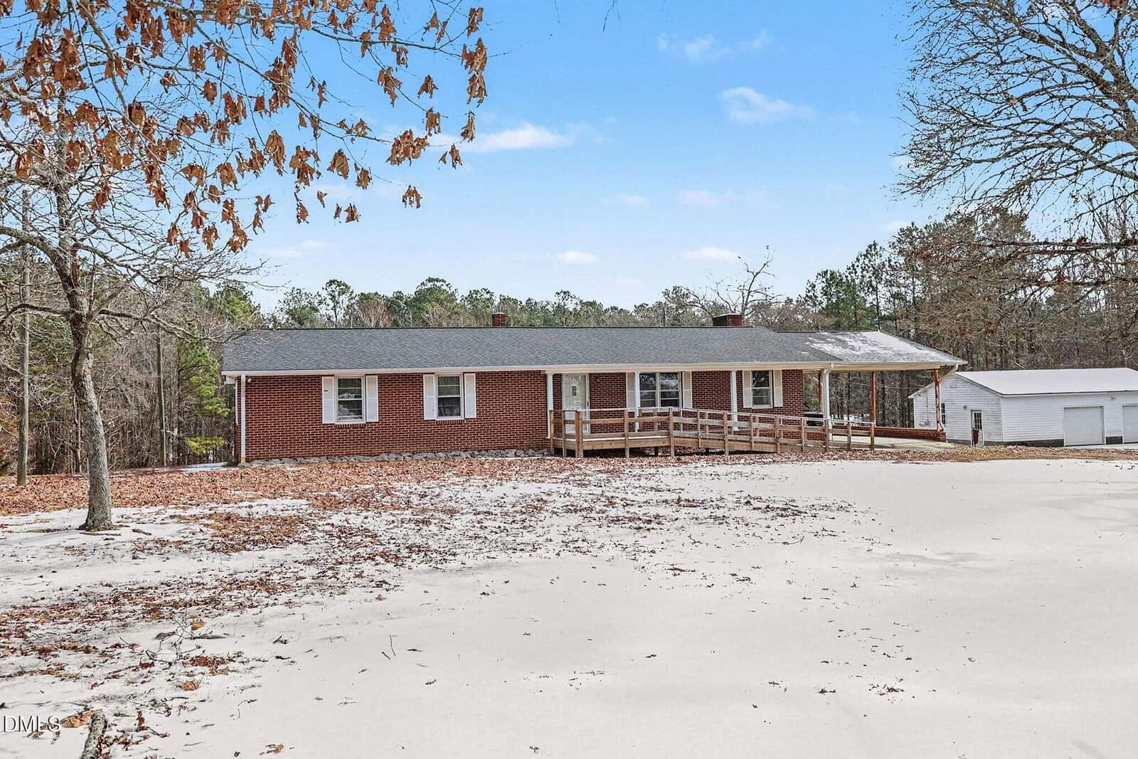 Single-story brick house with a gray roof, wooden wheelchair ramp, and large front yard with scattered leaves. Surrounded by trees, a detached white garage is visible on the right. Blue sky with light clouds overhead.