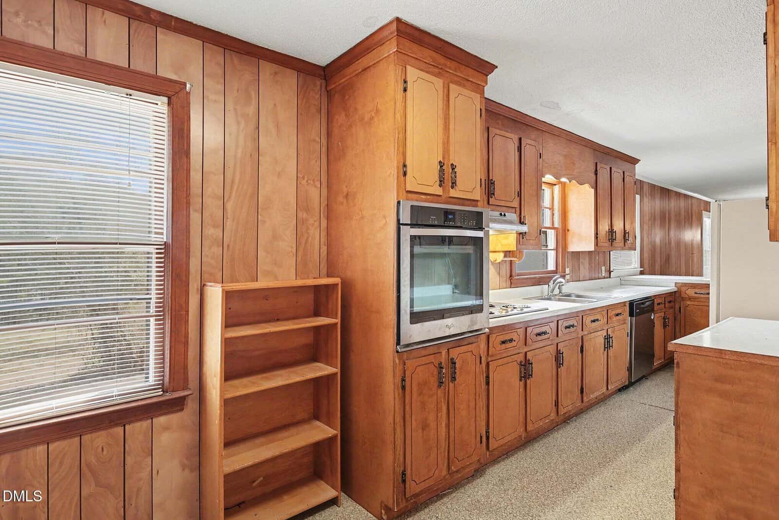 A vintage kitchen with wood-paneled walls, wooden cabinets, a built-in oven, electric stovetop, open shelving, and a window with blinds allowing sunlight into the room.