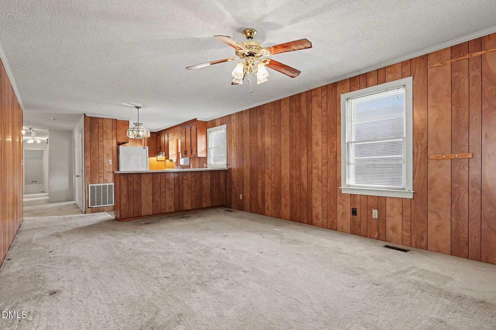 A living room with wood-paneled walls, beige carpet, a ceiling fan with lights, a window with blinds, and a partial view of the kitchen with wooden cabinets and a breakfast bar.