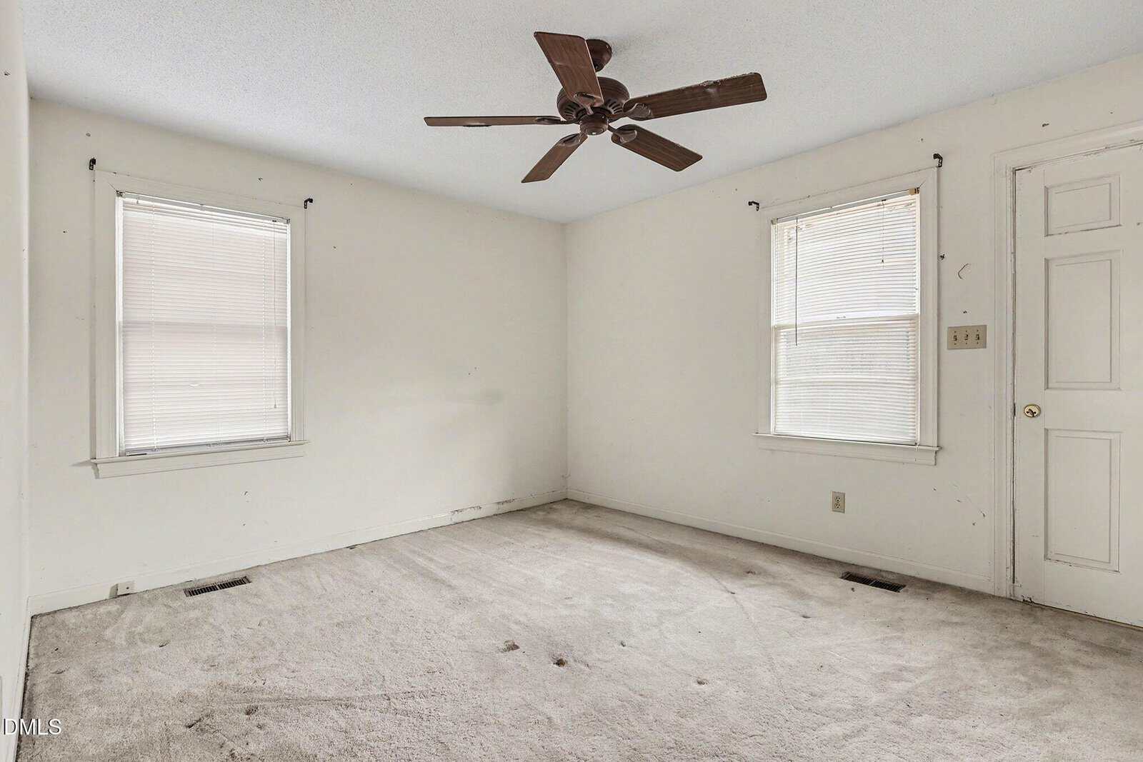 Empty room with two windows, a white door, beige carpet, white walls, and a brown ceiling fan. Natural light comes through the closed blinds on both windows. The room appears clean and undecorated.