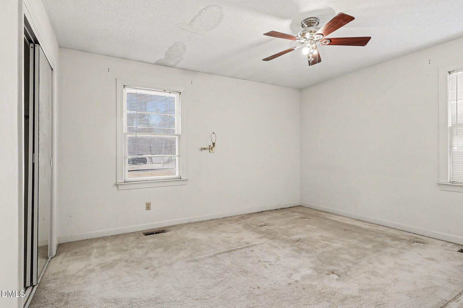 Empty room with two windows, beige carpet, white walls, and a ceiling fan with wooden blades. There are some stains on the ceiling and carpet, and a closet with sliding doors on the left.