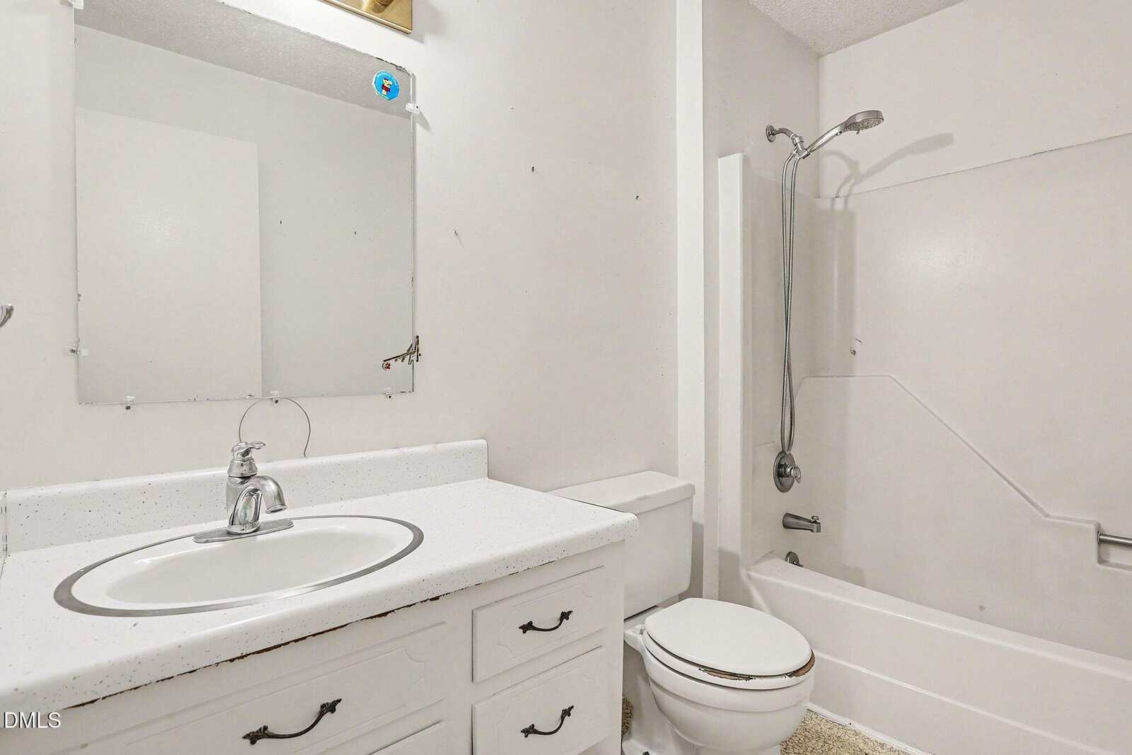 A simple white bathroom with a sink, countertop, mirror, toilet, and bathtub with a showerhead. The vanity has three drawers and cabinet doors below the sink. The walls and fixtures are all white.