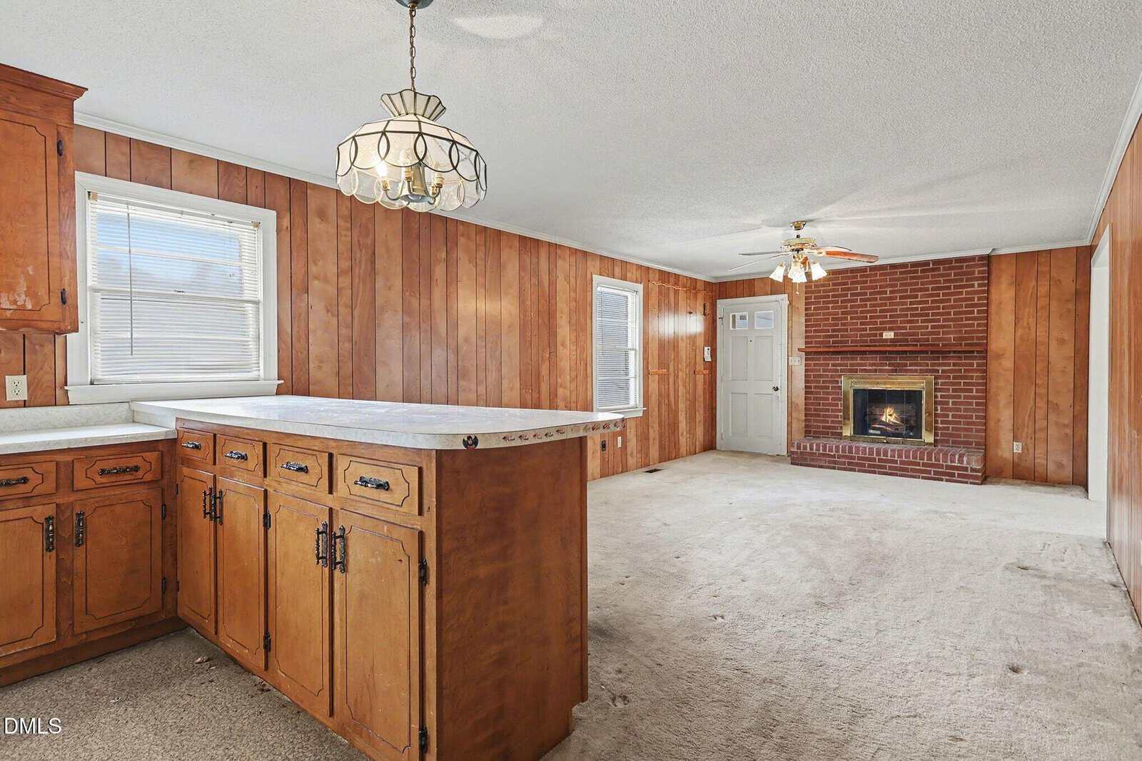 A living space with wood-paneled walls, beige carpet, a brick fireplace, ceiling fan, overhead light fixture, and a kitchen area with wooden cabinets and a white countertop.
