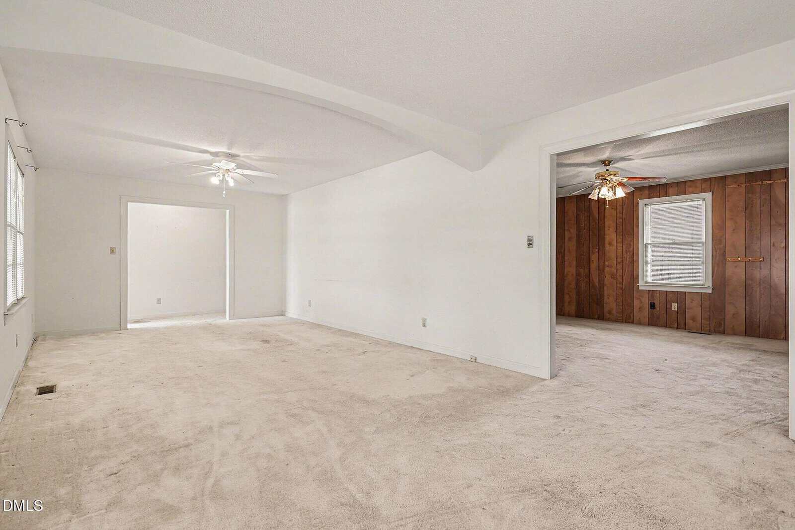 Spacious living room with light beige carpet, white walls, and ceiling fan; adjacent room has wood-paneled walls, carpet, two windows, and another ceiling fan. Both rooms appear empty and well-lit.