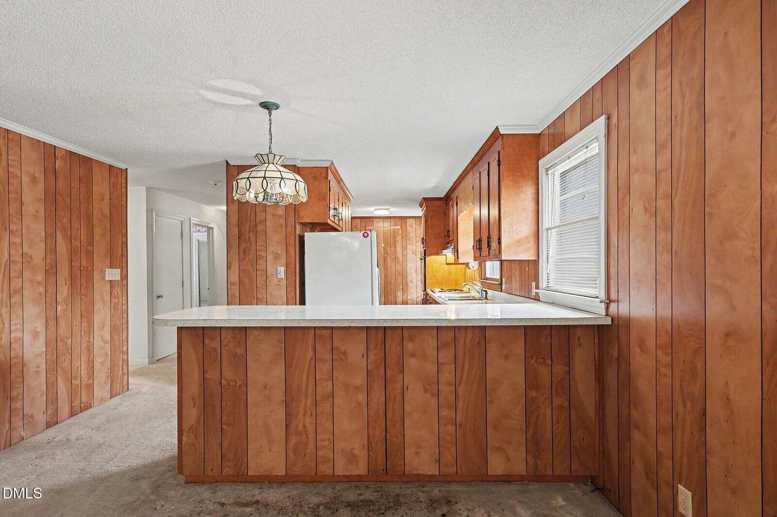 Kitchen with wood-paneled walls, a white countertop peninsula, brown cabinets, a white refrigerator, and a hanging glass light fixture. A window is above the counter, and a hallway is visible on the left.