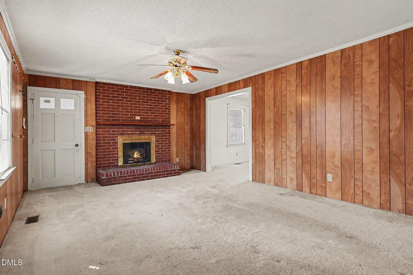 Living room with wood-paneled walls, beige carpet, brick fireplace with mantel, ceiling fan with lights, and a white front door with windows. An open doorway leads to another room.