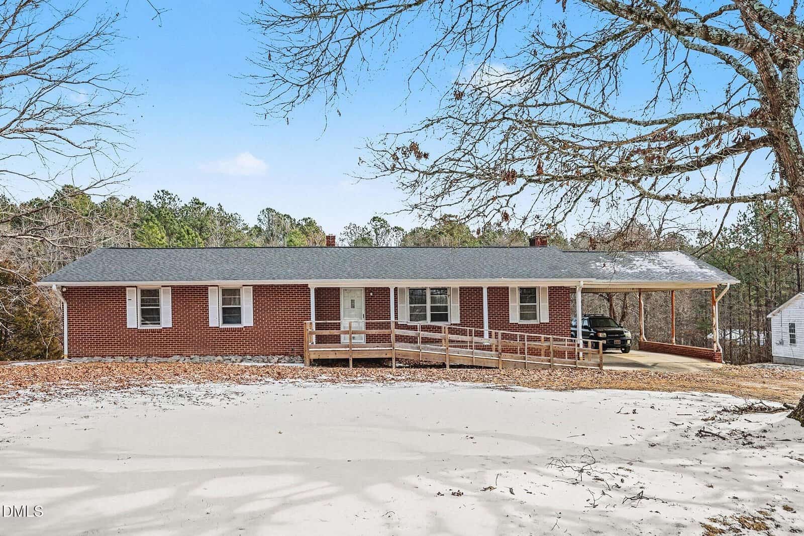 A single-story red brick house with a gray roof, front porch, and a wooden accessibility ramp. A carport is attached to the right, and trees surround the property. The yard has light patches of snow or sand.